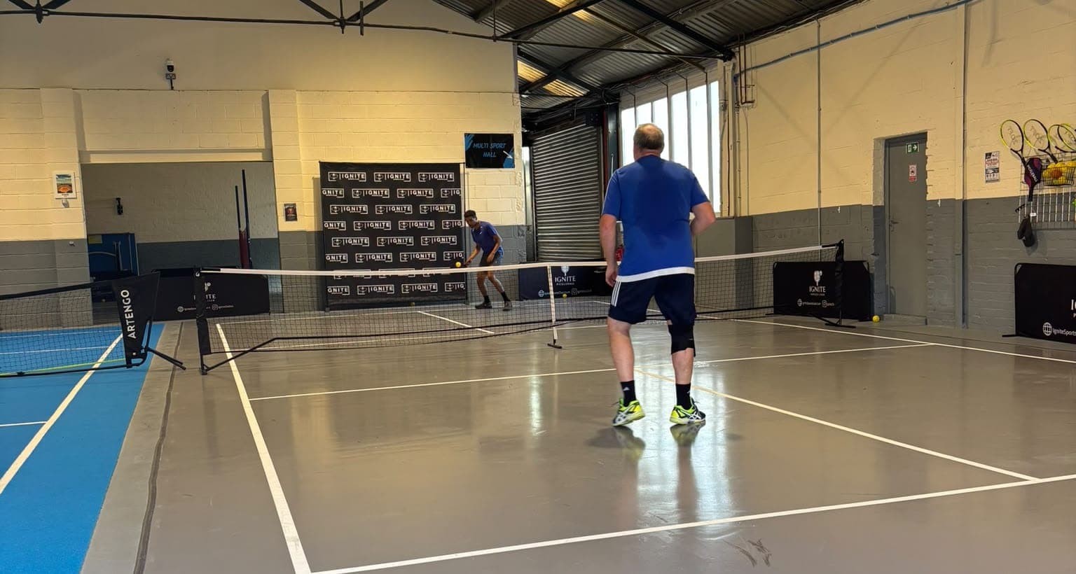 A pickleball rally on the grey court at Ignite, seen from the blue lounge area