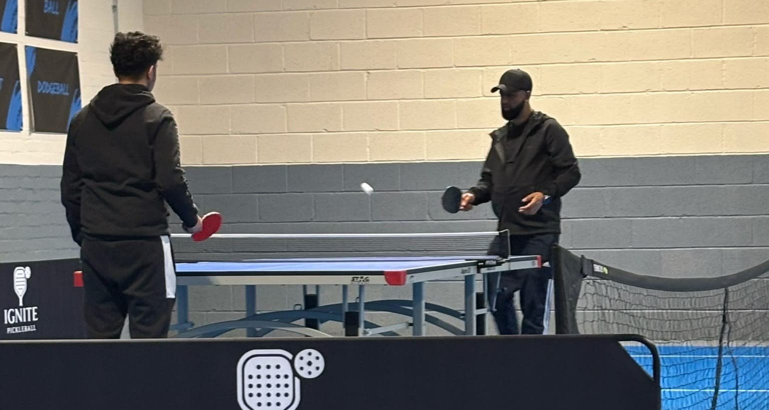 Two players at a table tennis table at Ignite, behind a black court banner