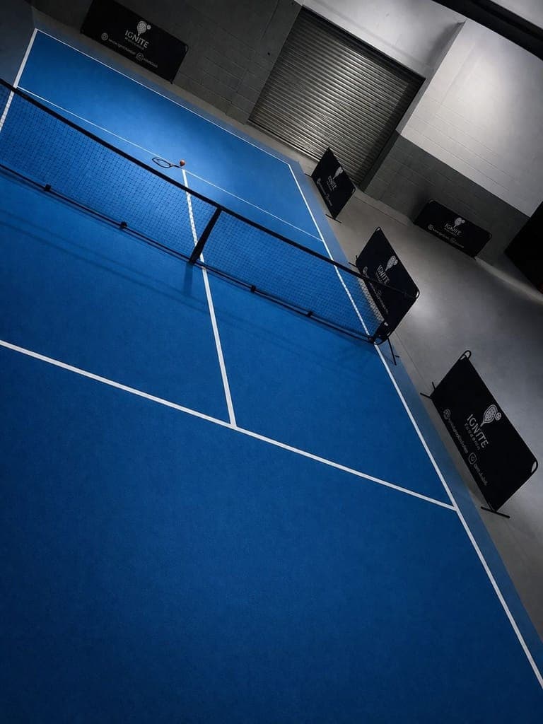 The blue carpet touch tennis court at Ignite, lit from above, with a racket and ball left by the net